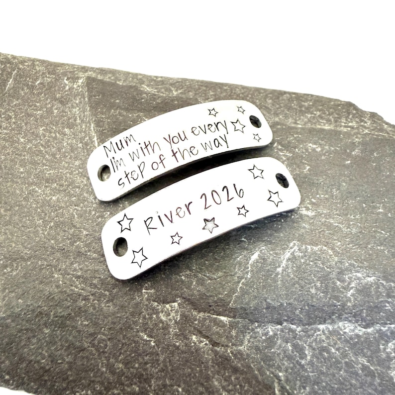 May include: Two silver-colored metal tags with rounded ends. One tag reads "Mum I'm with you every step of the way" and the other reads "River 2026". Both tags have star-shaped cutouts.