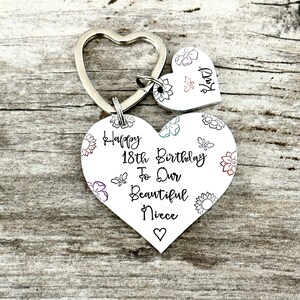 May include: A silver heart-shaped keychain with a smaller heart charm. The larger heart has a floral design and the text "Happy 18th Birthday To Our Beautiful Niece". The smaller heart charm has the text "Katy".