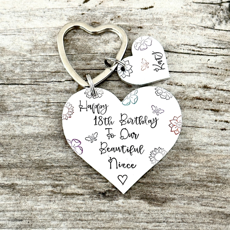 May include: A silver heart-shaped keychain with a smaller heart charm. The larger heart has a floral design and the text "Happy 18th Birthday To Our Beautiful Niece". The smaller heart charm has the text "Katy".