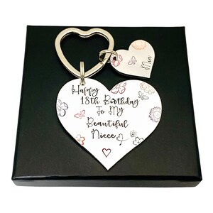 May include: A white heart-shaped keychain with the text "Happy 18th Birthday To My Beautiful Niece" and a small heart at the bottom. The keychain is attached to a silver key ring with a smaller heart-shaped charm that says "Niece". The keychain is in a black gift box.
