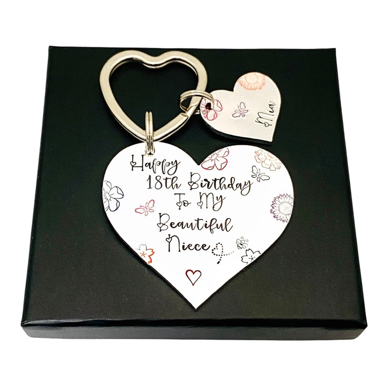 May include: A white heart-shaped keychain with the text "Happy 18th Birthday To My Beautiful Niece" and a small heart at the bottom. The keychain is attached to a silver key ring with a smaller heart-shaped charm that says "Niece". The keychain is in a black gift box.