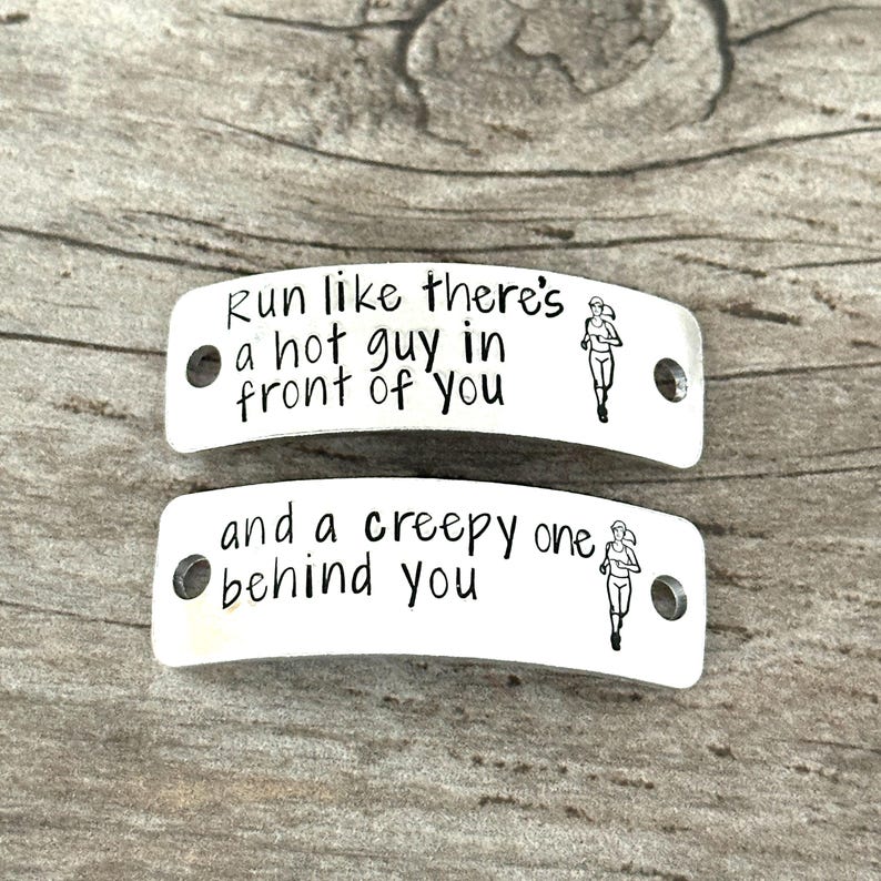 May include: Two curved silver-toned metal shoe tags with holes on each end. The top tag features the black text "Run like there's a hot guy in front of you" and a runner icon. The bottom tag features the text "and a creepy one behind you".