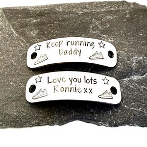 May include: Two curved, white, rectangular metal shoe tags with black engraved text and graphics. The top tag reads "Keep running Daddy" with star and sneaker icons. The bottom tag reads "Love you lots Ronnie xx" with star and sneaker icons.