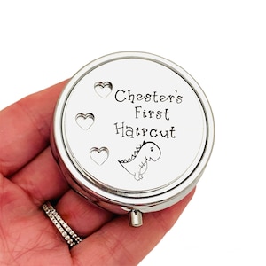 May include: A round, silver-colored pill box with the text "Chester's First Haircut" and a cartoon dinosaur illustration. The box has three heart-shaped designs. A small, silver button is on the side.