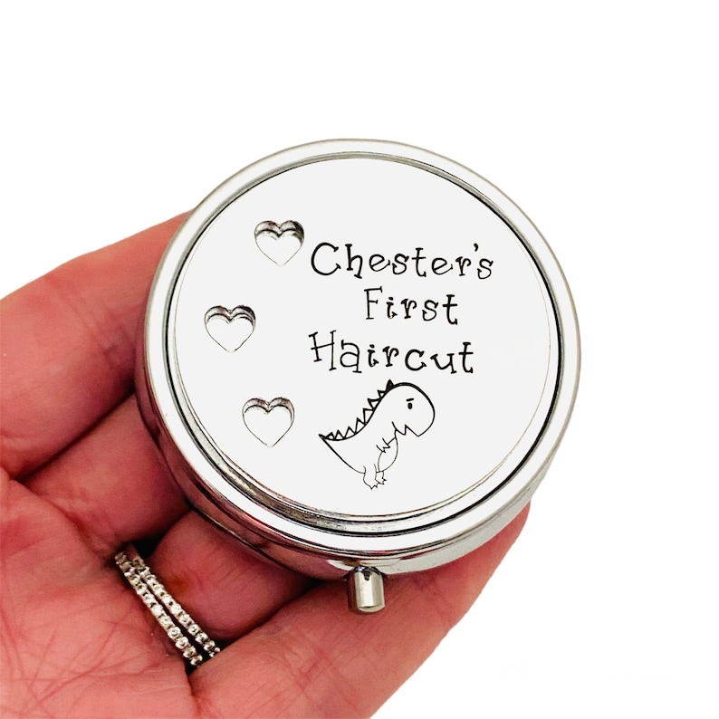 May include: A round, silver-colored pill box with the text "Chester's First Haircut" and a cartoon dinosaur illustration. The box has three heart-shaped designs. A small, silver button is on the side.