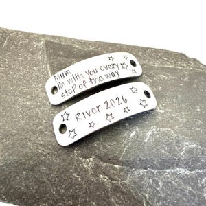 May include: Two curved silver-toned metal tags with holes at each end. The top tag is engraved with the text "Mum I'm with you every step of the way" and small stars. The bottom tag is engraved with the text "River 2026" and several star outlines.