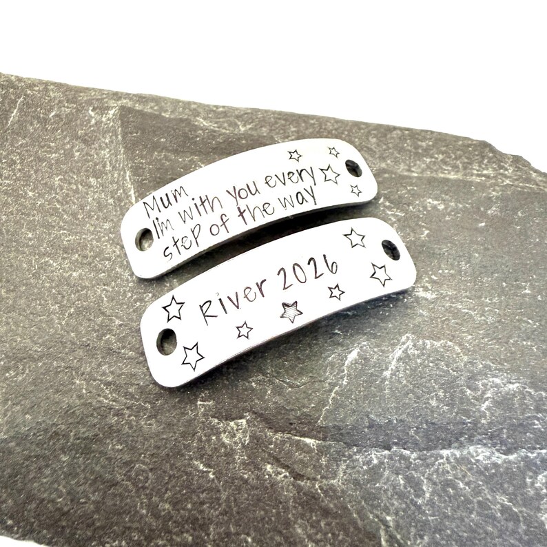 May include: Two curved silver-toned metal tags with holes at each end. The top tag is engraved with the text "Mum I'm with you every step of the way" and small stars. The bottom tag is engraved with the text "River 2026" and several star outlines.
