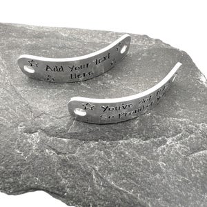 May include: Two curved silver-toned metal tags with holes at each end. One tag features the engraved text "Add Your Text Here" and the other features "You've got this! So proud of you!" Both tags are decorated with small engraved star shapes.