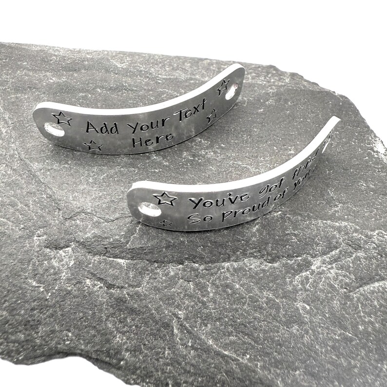 May include: Two curved silver-toned metal tags with holes at each end. One tag features the engraved text "Add Your Text Here" and the other features "You've got this! So proud of you!" Both tags are decorated with small engraved star shapes.