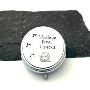 May include: A silver-colored, round keepsake box with the text "Wesley's First Haircut" and a train illustration. Three star cutouts are above the text. The box has a small, silver-colored handle.