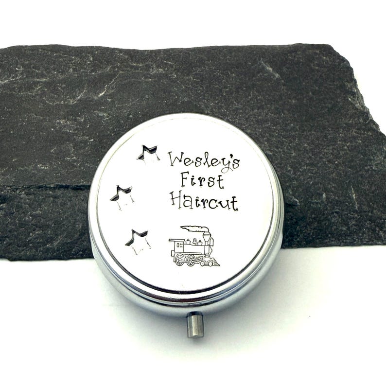 May include: A silver-colored, round keepsake box with the text "Wesley's First Haircut" and a train illustration. Three star cutouts are above the text. The box has a small, silver-colored handle.