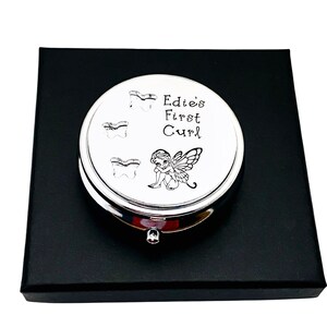 May include: A silver-colored, round keepsake box with the text "Edie's First Curl" and three tooth illustrations. A small fairy illustration is also present. The box is presented on a black box.