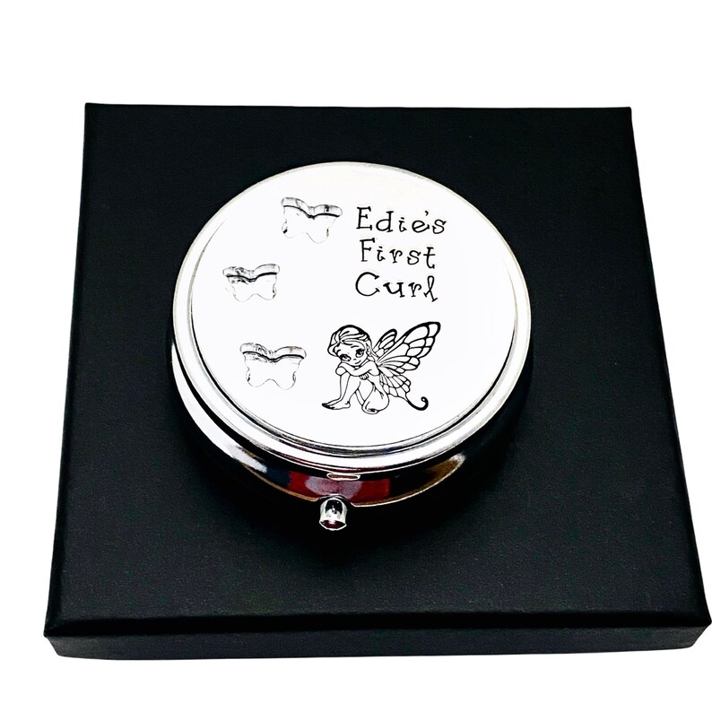 May include: A silver-colored, round keepsake box with the text "Edie's First Curl" and three tooth illustrations. A small fairy illustration is also present. The box is presented on a black box.