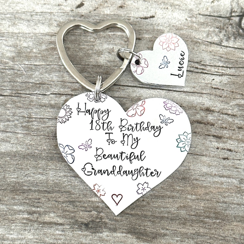 May include: Silver heart-shaped keychain with a message that reads "Happy 18th Birthday To My Beautiful Granddaughter" with floral designs. A smaller heart-shaped tag with the name "Lucille" is attached to the keychain.