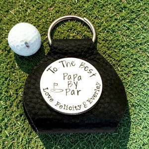 May include: A black leather golf keychain with a silver ring and a white circular insert. The insert reads "To The Best Papa By Par Love Felicity & Everley" around a golf course graphic. A white golf ball rests on the green grass.