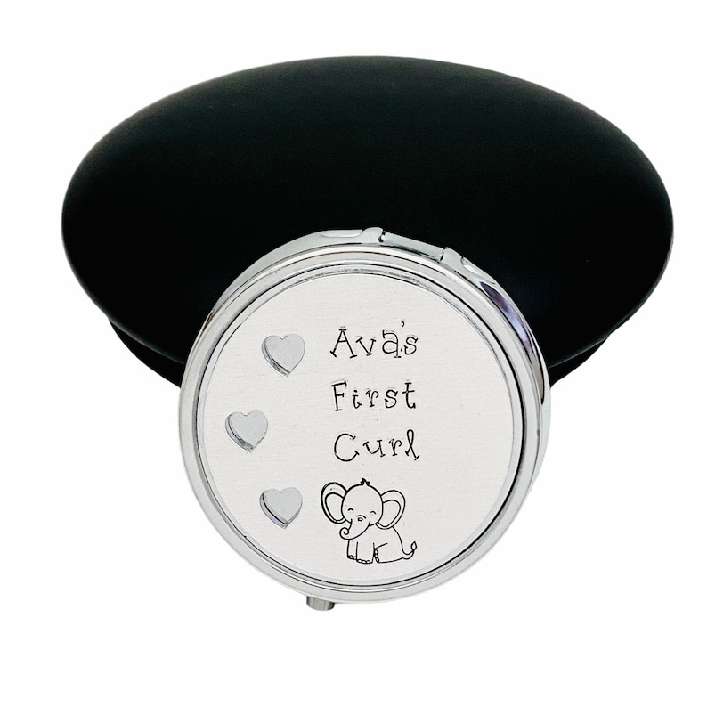 May include: A silver compact mirror with the text "Ava's First Curl" and a small elephant illustration. The mirror has three heart designs and a black, oval-shaped case.