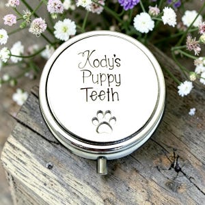 May include: A round, silver-colored metal keepsake tin with a lid featuring the black printed text Kody's Puppy Teeth and a small paw print icon. The container has a smooth, reflective surface and a small metal latch on the front edge.