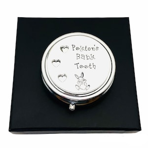 Puede incluir: Caja plateada para guardar dientes de beb&eacute; con el texto "Peyton's Baby Teeth" y una ilustraci&oacute;n de un conejito. La caja redonda se presenta sobre una caja negra. Para guardar dientes de leche.
