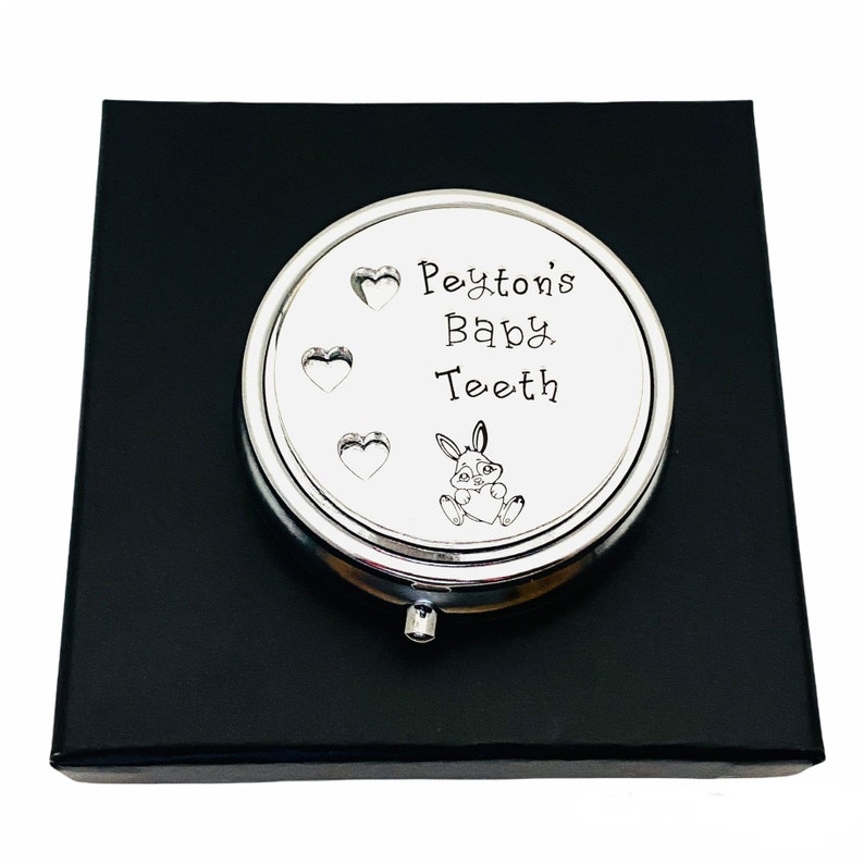 Puede incluir: Caja plateada para guardar dientes de beb&eacute; con el texto "Peyton's Baby Teeth" y una ilustraci&oacute;n de un conejito. La caja redonda se presenta sobre una caja negra. Para guardar dientes de leche.