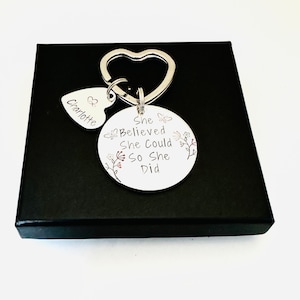 Pode incluir: Um porta-chaves de metal prateado com um anel em forma de cora&ccedil;&atilde;o, um pequeno pendente em forma de cora&ccedil;&atilde;o gravado com o nome Charlotte e um disco circular maior gravado com o texto She Believed She Could So She Did, decorado com motivos florais e borboletas.