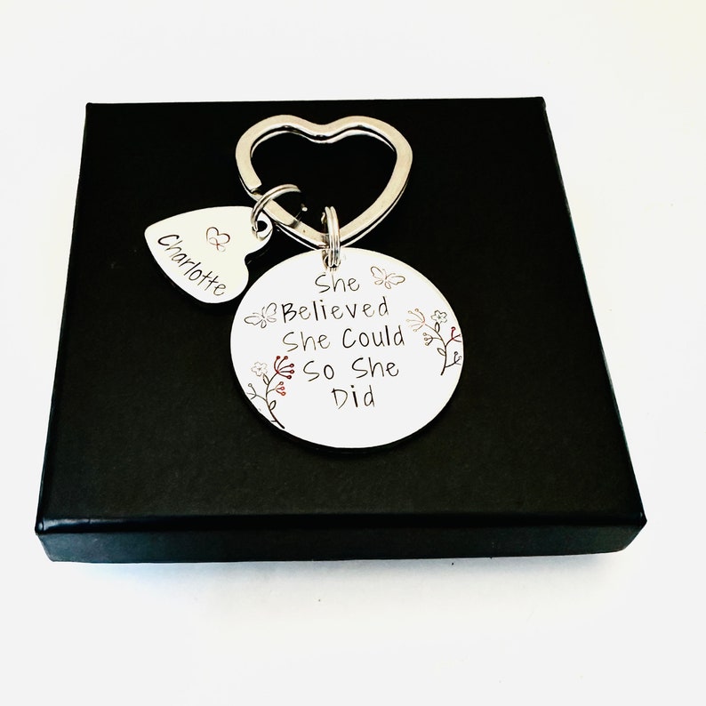 Pode incluir: Um porta-chaves de metal prateado com um anel em forma de cora&ccedil;&atilde;o, um pequeno pendente em forma de cora&ccedil;&atilde;o gravado com o nome Charlotte e um disco circular maior gravado com o texto She Believed She Could So She Did, decorado com motivos florais e borboletas.