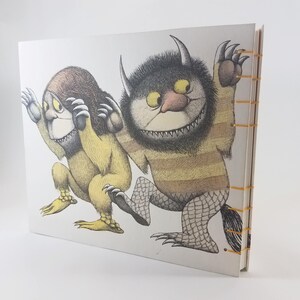 Może przedstawiać: Ilustracja okładki książki przedstawiająca dwie futrzane istoty. Jedna to ż&oacute;łto-brązowe stworzenie z dużymi uszami i szerokimi ustami. Druga to czarno-brązowe stworzenie z paskowanym ciałem i szerokim, zębatym uśmiechem. Okładka książki jest biała z ż&oacute;łtym oprawieniem.
