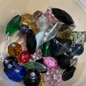 May include: A collection of colorful glass beads in various shapes and sizes, including round, oval, and faceted. The beads are a mix of green, blue, red, pink, brown, and clear glass. Some beads have silver accents.