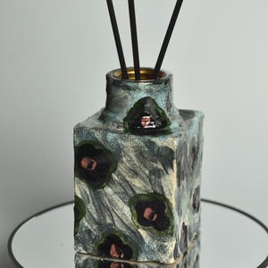 May include: A decorative ceramic diffuser bottle with a square shape and a textured, crackled finish. The bottle is painted in shades of blue and gray, with dark green and pink floral accents. Black reeds are inserted into the bottle.