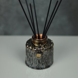 May include: Black ceramic diffuser bottle with a gold accent and a copper top. The bottle has a textured surface and is filled with black diffuser sticks.