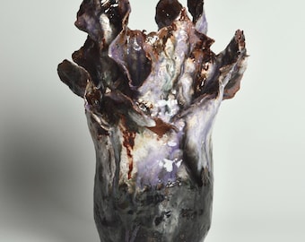 May include: A ceramic vase with a unique, organic shape and a textured, multi-colored glaze. The vase is dark brown, purple, and white with a mottled appearance.