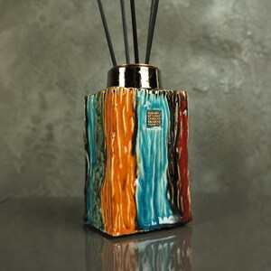 May include: A ceramic diffuser with a square shape and a colorful design. The diffuser has a blue, orange, and green striped pattern with a gold accent. It is filled with black sticks.