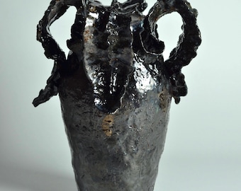 May include: A dark, glossy, charcoal-colored ceramic vase with a highly textured, uneven surface. The vessel features two prominent, ruffled, leaf-like handles extending from the sides and a wide, irregular opening at the top.