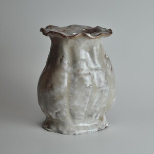 May include: A white ceramic vase with a textured, organic shape and a scalloped rim. The vase has a crackled glaze and a brown tint.