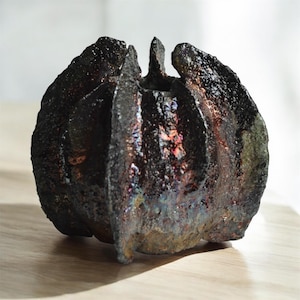 May include: A dark, textured ceramic vase with a unique, organic shape. The vase has a rough, almost volcanic surface with iridescent highlights of red, purple, and green. The design features vertical ridges and a central opening, suggesting a sculptural art piece.