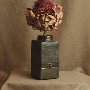 May include: A dried artichoke flower in a dark green and brown ceramic vase with a gold band. The vase is sitting on a brown fabric surface.