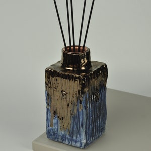 May include: A square ceramic diffuser bottle with a textured, blue and gold glaze. Black diffuser sticks are inserted into the bottle.