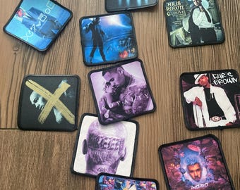 May include: A collection of square fabric patches with various designs. The patches feature images of musicians, album art, and text. The color palette includes blues, purples, and black, with some patches displaying text such as "Royalty" and "Indigo".