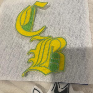 Könnte beinhalten: Gelb und grün gestickte Buchstaben "CB" in einem gotischen Schriftstil. Die Buchstaben befinden sich auf einem weißen Filzhintergrund. Zusätzliche weiße Filzaufnäher mit schwarzen Umrissen sind sichtbar.
