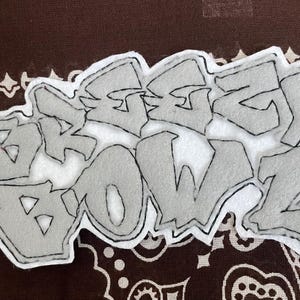 Może przedstawiać: Szara filcowa naszywka z napisem "DREAZ BOWZ" w stylu graffiti, obrysowana na czarno. Naszywka znajduje się na brązowo-białym wzorzystym tle.