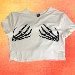 May include: White cropped t-shirt with a black skeleton hand graphic on the front. The shirt has short sleeves and a crew neck.