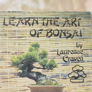 May include: A book titled "Learn the Art of Bonsai" by Laurence Craven, featuring a photo of a bonsai tree in a brown pot on a wooden stand. The background is a bamboo mat.