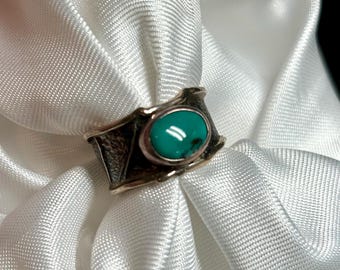 Puede incluir: Primer plano de un anillo con una piedra turquesa. El anillo tiene una banda de metal oscuro texturizado con forma rectangular. La turquesa ovalada está engastada en un bisel dorado.