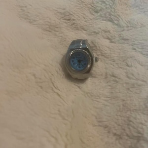 May include: A silver ring watch with a light blue face. The watch features a classic design with a round face and a band made of small, connected metal pieces. The watch face displays numbers and hands.