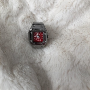 May include: A silver quartz ring watch with a square red face. The watch has a silver band and a small crown. The watch face displays the numbers 1 to 12, with the brand name "BAZHI" at the top.