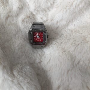 May include: A silver watch with a red face and black hands. The watch has a square face and a silver band. The watch is on a white furry surface.