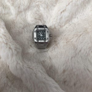 May include: A silver ring watch with a square black face. The watch has a quartz movement and displays the time with white numerals and hands. The ring watch is set against a fluffy, off-white background.
