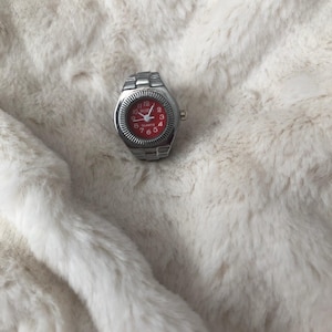 May include: A silver watch with a red face and silver hands. The watch has a silver band and a quartz movement.