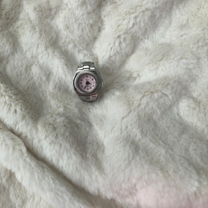 May include: A silver ring watch with a pink face and black numerals. The watch has a classic design with a round face and a metal band. The watch is set against a white, fluffy background.