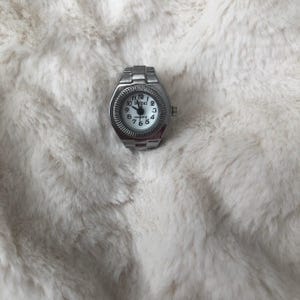 May include: A silver ring watch with a white face and black numerals. The watch has a quartz movement and the brand name "BOZHI" is printed on the face. The watch is set against a fluffy, white background.