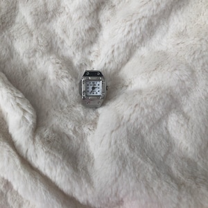 May include: A small, silver wristwatch with a square face and a white dial. The watch has black numerals and hands, and is set against a fluffy, white background. The watch face reads "BOOM 2".